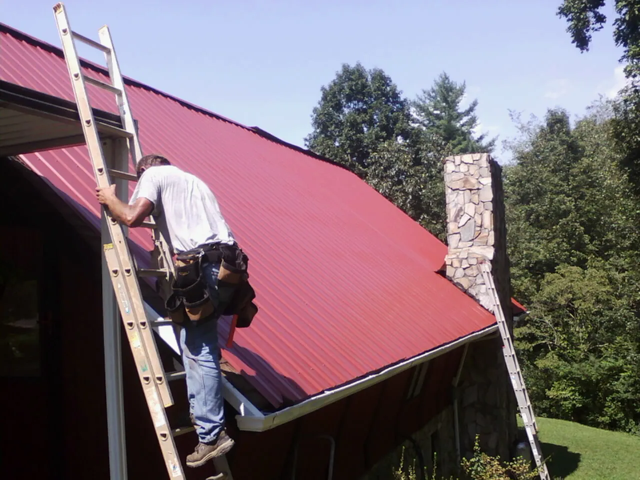 Roofer on a ladder beside a red metal roof and stone chimney — Soffit & Fascia Repair by Roofing Brothers in Tygh Valley